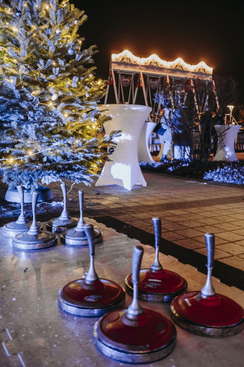 Vordergründig ist der Ansatz einer Eisstockbahn zu sehen. Links im Anschnitt ist ein Weihnachtsbaum zu sehen und im Hintergrund eine Schiffsschaukel.